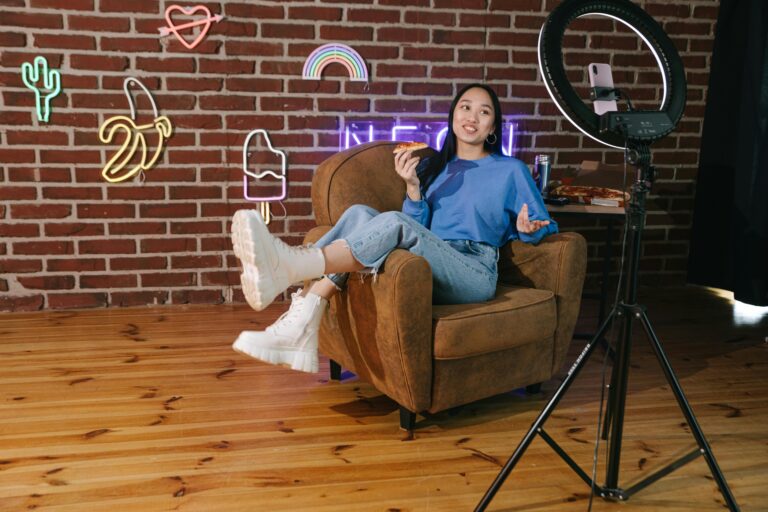 Young Asian woman influencer streaming live video while enjoying pizza in a stylish room with neon décor.